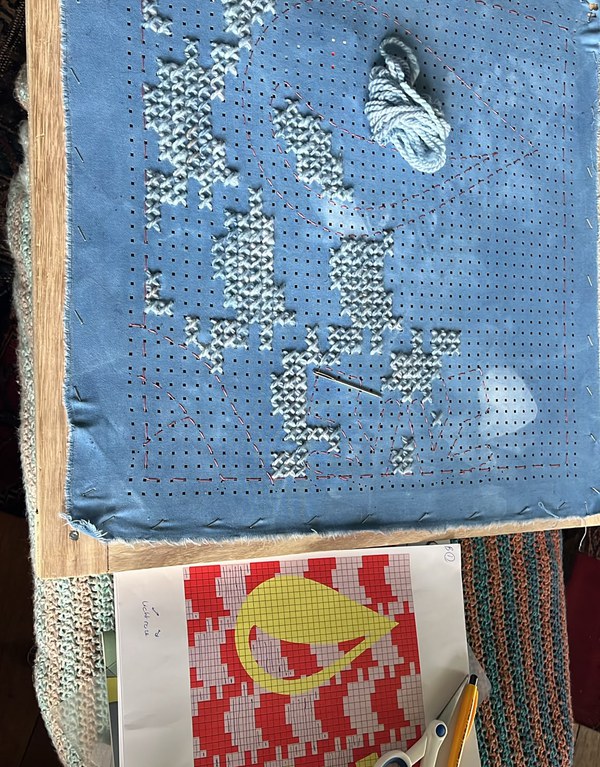 Een half afgeborduurd paneel van lichtblauwe stof in een houten borduurraam, met daaronder het patroonblad met het ontwerp.