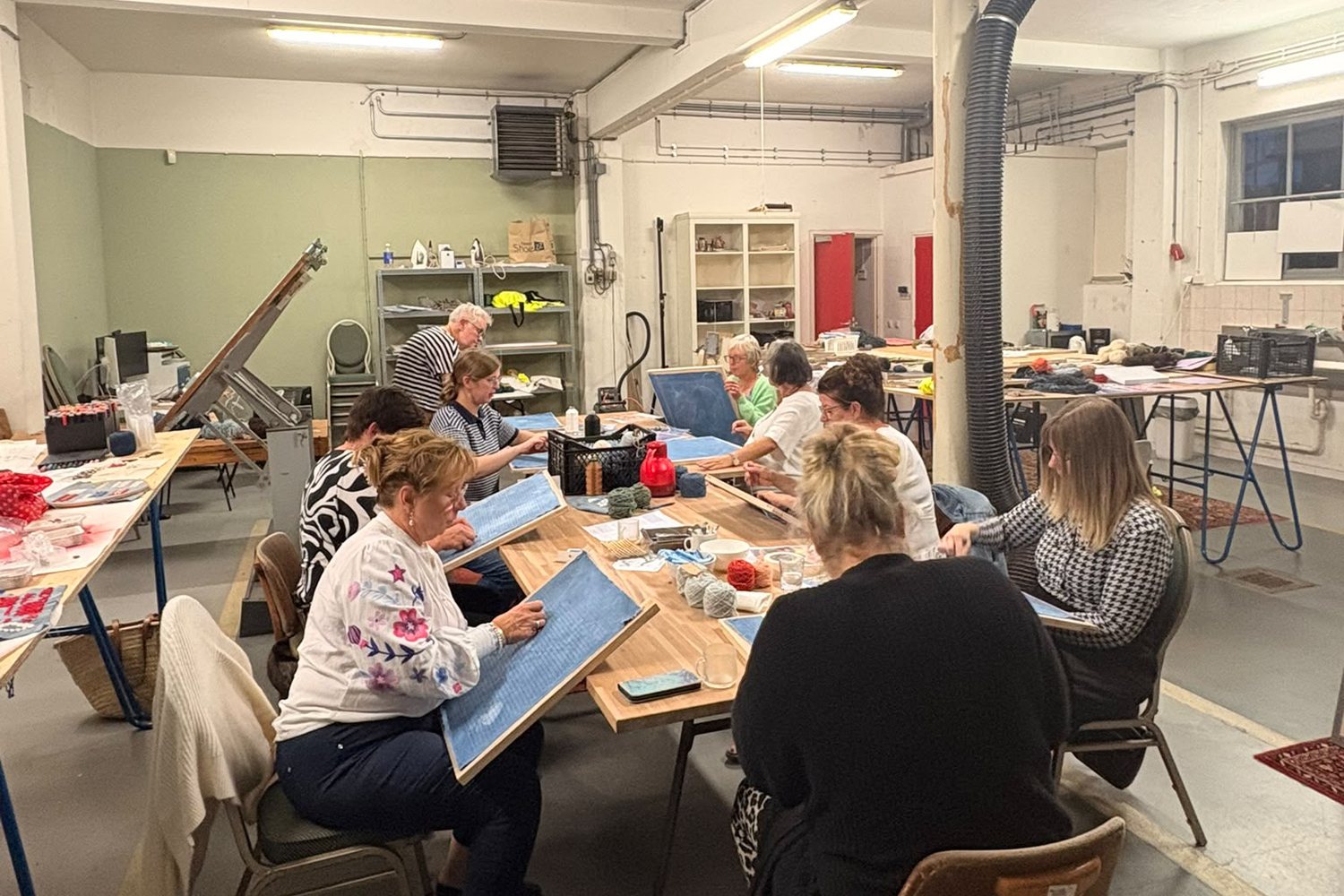 Een groep vrouwen borduurt aan tafel in het atelier van de Cultuurschuur, elk met een borduurraam met lichtblauwe stof.